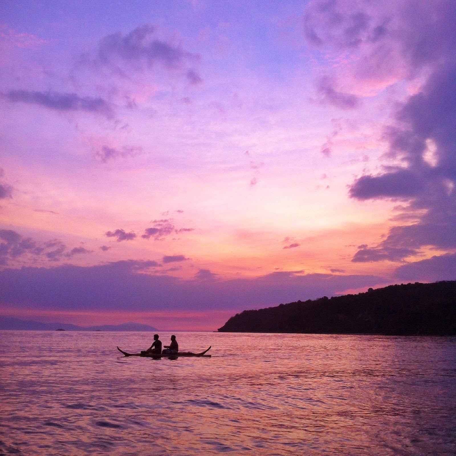 Silhouette of two people in a narrow outrigger boat on calm water at sunset, soft purple and pink sky over a dark headland, quiet and reflective mood.