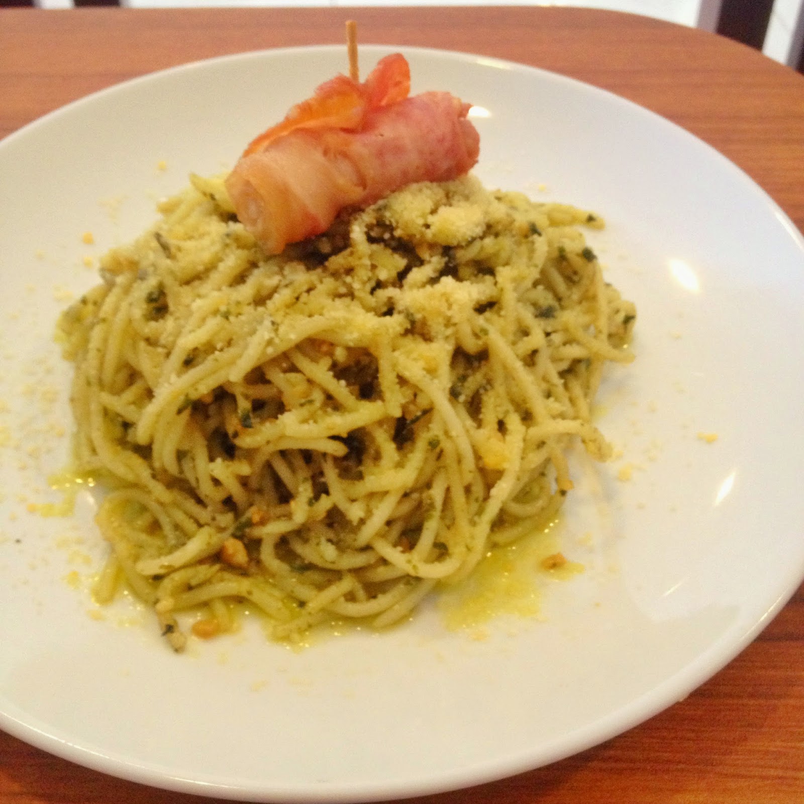 Close-up of a plate of pesto spaghetti topped with grated cheese and a rolled slice of cured meat on a skewer, placed on a wooden board with soft natural daylight and warm green and cream tones