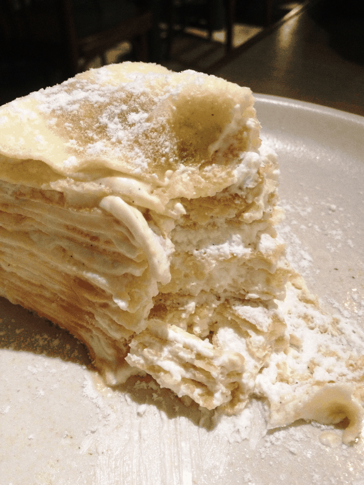 Close-up of a sliced layered crepe cake with cream filling and powdered sugar dusting on a plate, pale yellow and white tones with a soft, calm mood