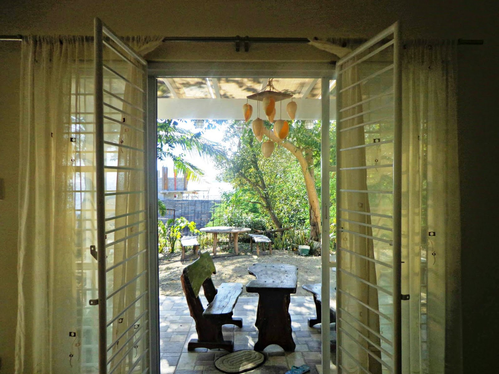 View through open doors to an outdoor patio with a wooden table and benches, surrounded by greenery, soft natural daylight, calm and homey atmosphere.